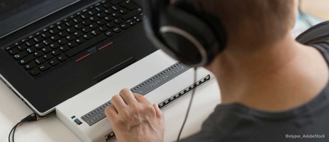 Assistive Technologie bei barrierefreien PDF: Ein Man hat Kopfhörer auf und sitzt vor einem Laptop und verwendet dazu ein Braillezeilen-Gerät.